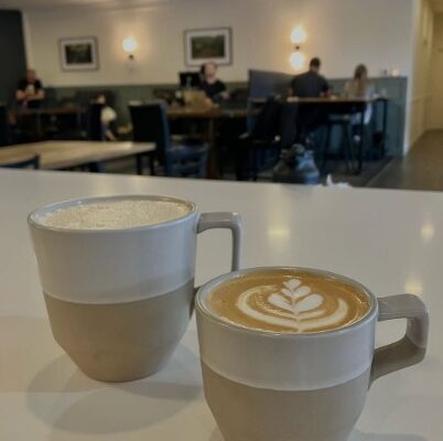 Two coffee drinks sitting on the counter with a view of cafe seating in the background.